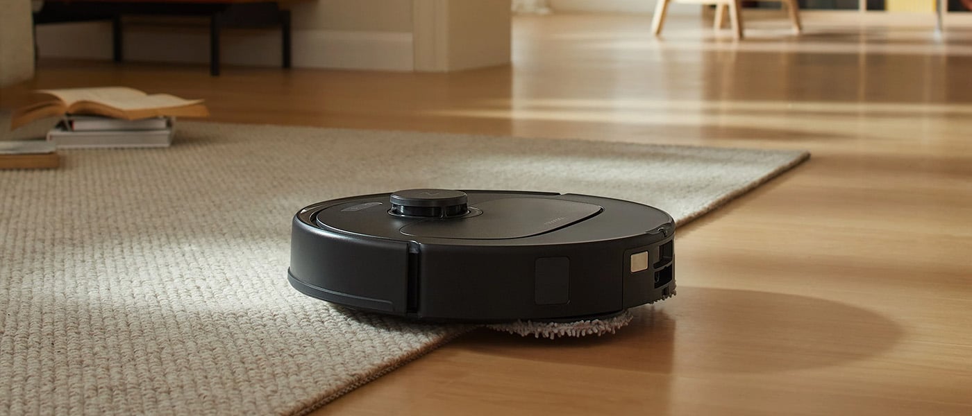 Svart robotdammsugare i drift, kör från ljus trämatta upp på en beige matta i ett ljust vardagsrum.