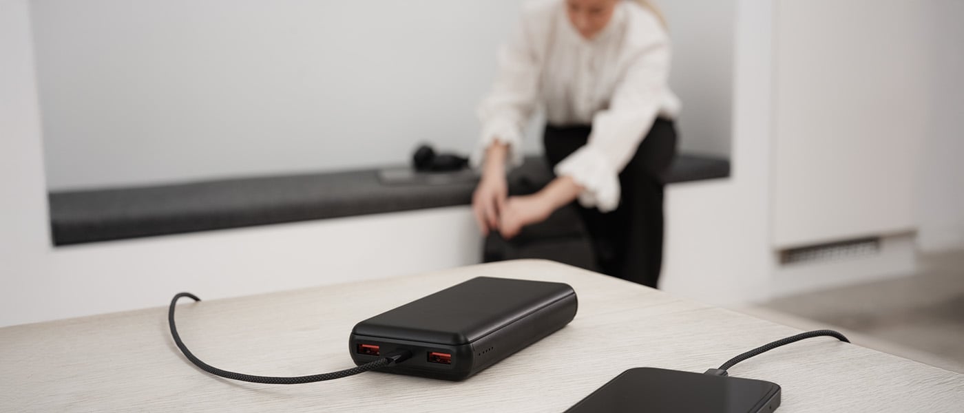 Svart powerbank med to USB-porter lader en smarttelefon via kabel på et lyst bord, uskarp person i bakgrunnen.