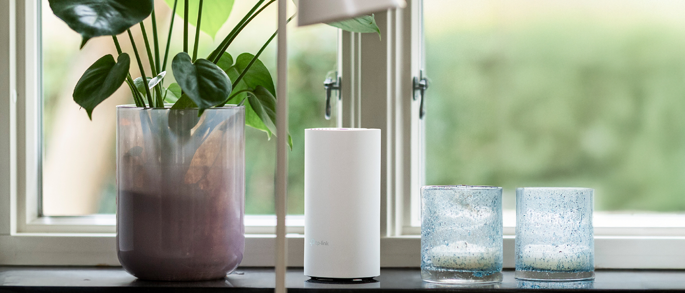 Hvit TP-Link-ruter står i vinduskarmen mellom en grønn plante i lilla potte og to blå telysholdere i glass.