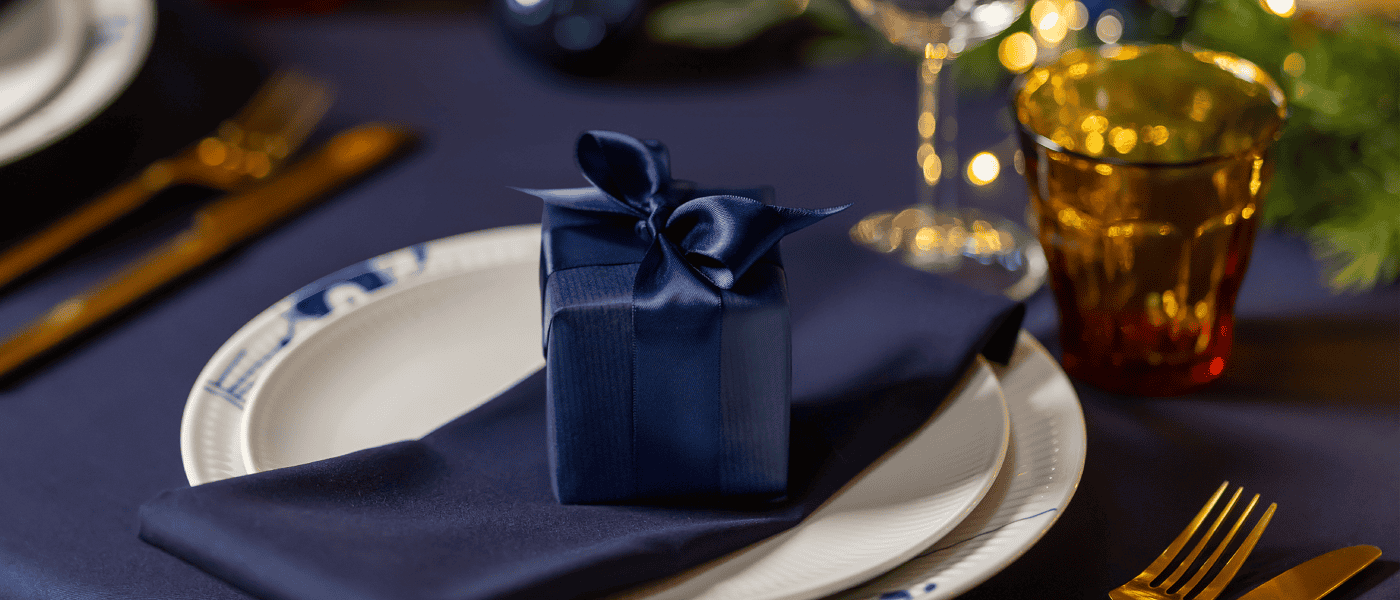 Blå gaveeske med sløyfe plassert på en mørk serviett på en juledekket tallerken med gullbestikk og glass.