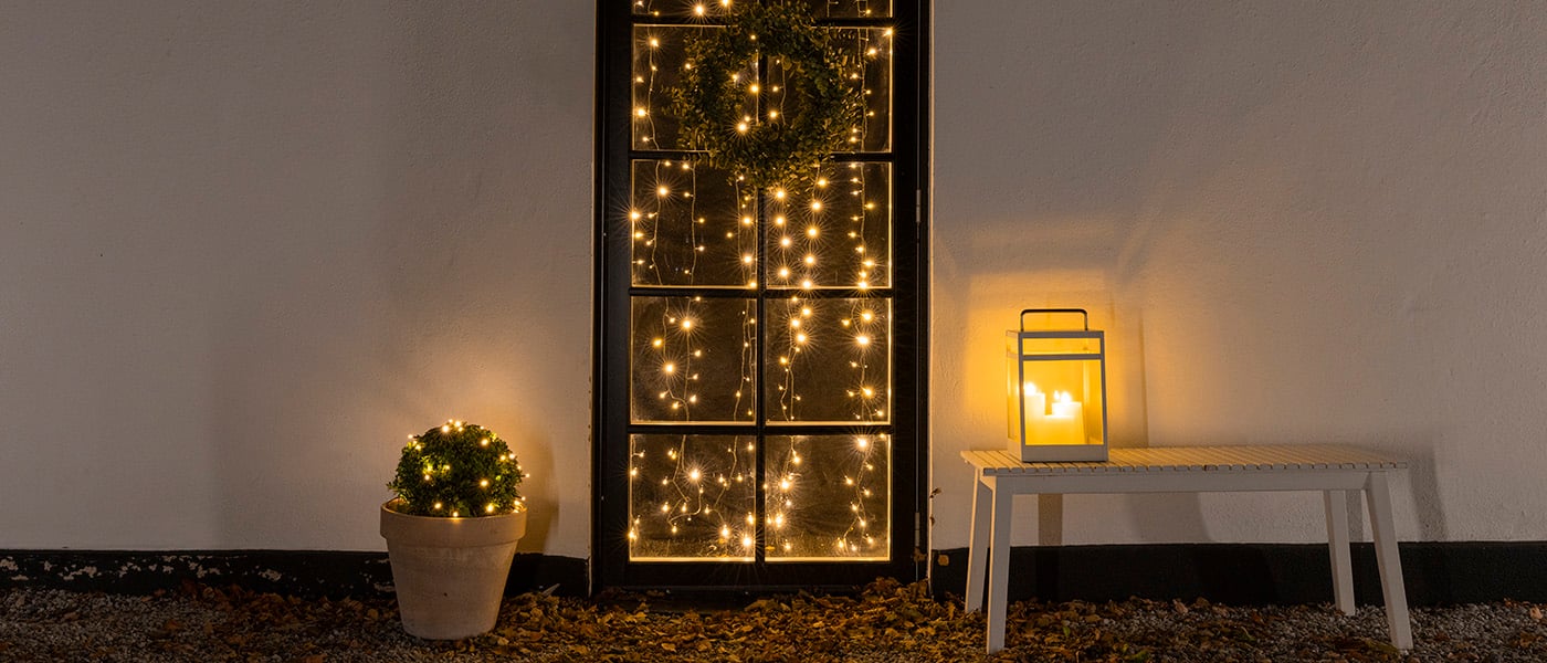 Svart dörr med ljusslingor och grön krans, kruka med ljusslingor till vänster och lykta med LED-ljus på vit bänk till höger.