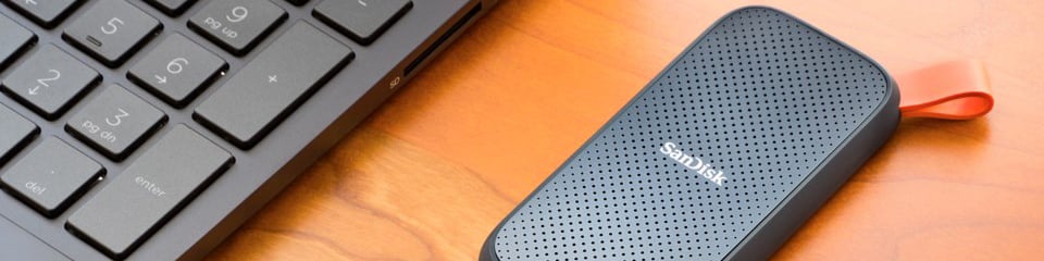 SanDisk extern SSD i svart med röd ögla, placerad bredvid en bärbar dators tangentbord på ett träbord.