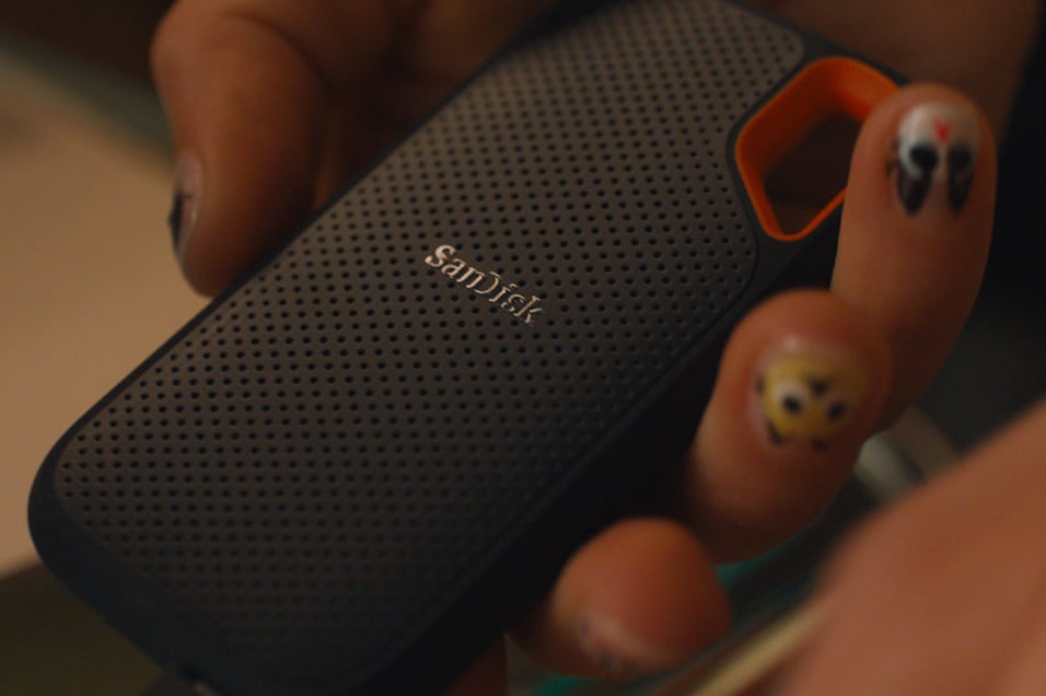 Bärbar SanDisk SSD i svart med orange fäste och strukturerad yta, hålls i handen. Logotypen “SanDisk” syns på framsidan.