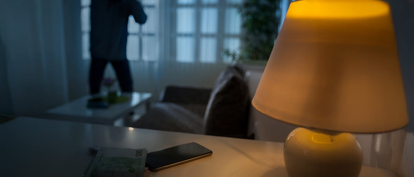 Silhuett av en person i mörk luvtröja vid ett fönster i kvällsbelysning, med en tänd gul lampskärm i förgrunden.