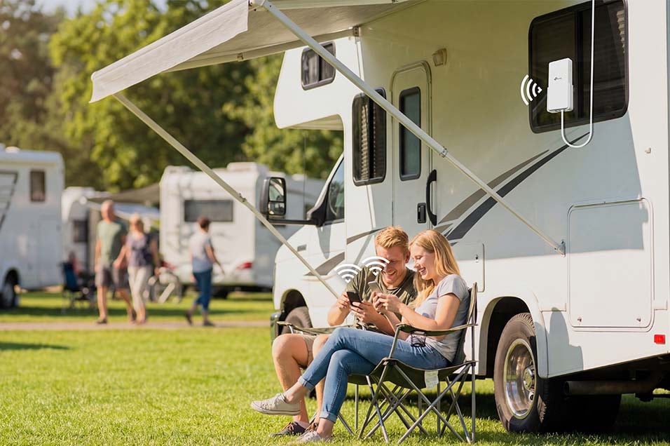Par använder smartphone utanför en husbil på campingplats med TP-Link Archer utomhusrouter monterad på väggen.