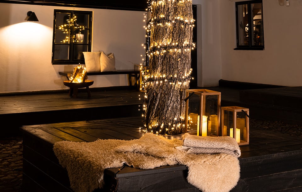 Opplyst trestamme med lyslenke, tre lykter med LED-lys og tepper på en terrasse i kveldsbelysning.