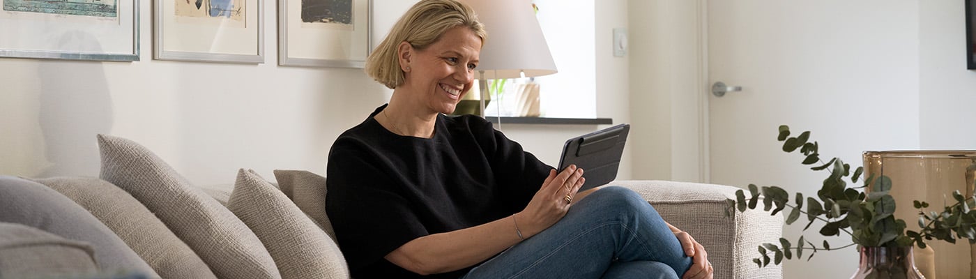 Smilende kvinne sitter i sofa og bruker et nettbrett i en lys stue med moderne innredning.