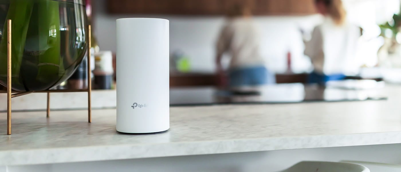 Vit TP-Link-router placerad på köksbänk med suddig bakgrund där två personer syns i ett modernt kök.