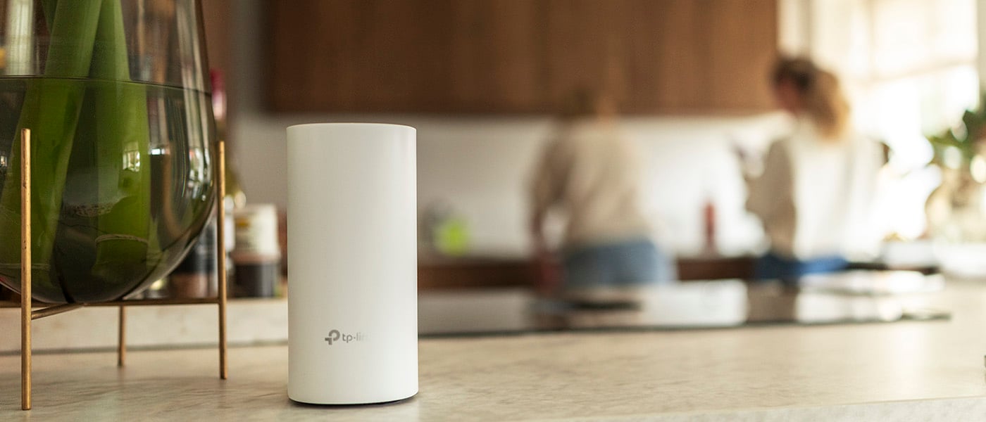 Vit TP-Link-router placerad på köksbänk med suddig bakgrund där två personer syns i ett modernt kök.