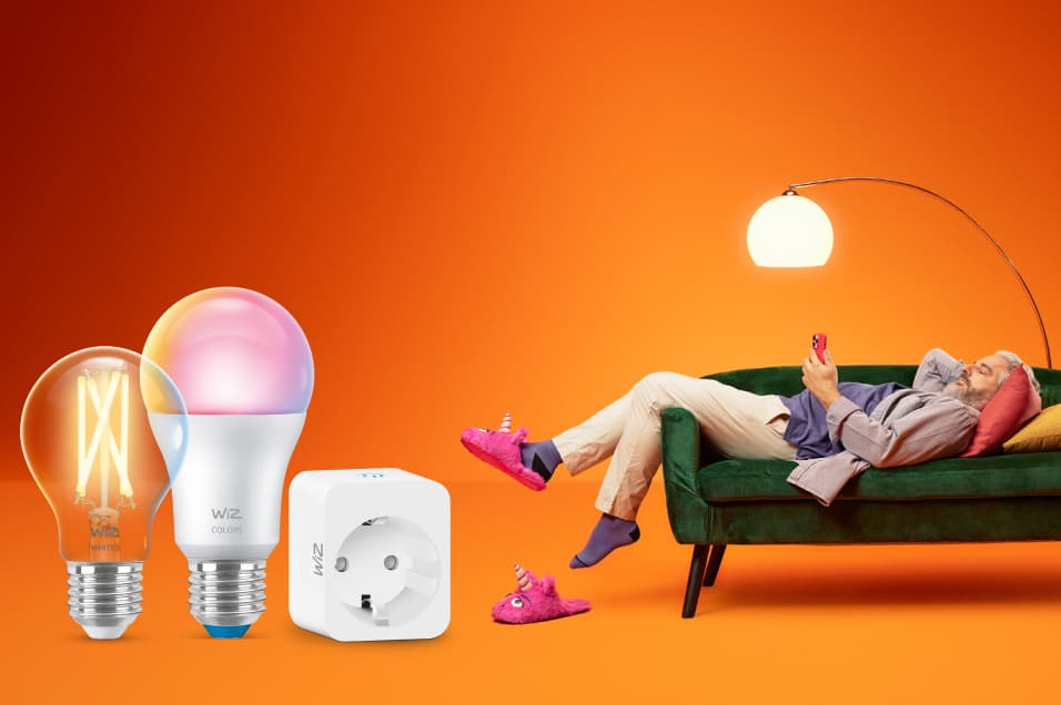 Smart belysning från WiZ, inklusive LED-lampor, ljusslinga och kontakt, med avslappnad man på grön soffa i orange miljö.