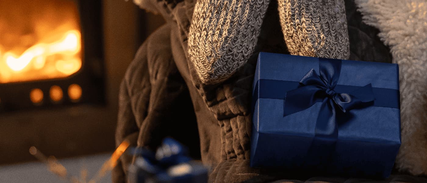 Mörkblå present med sidenband framför stickade sockor vid en öppen brasa i varm vintermiljö.