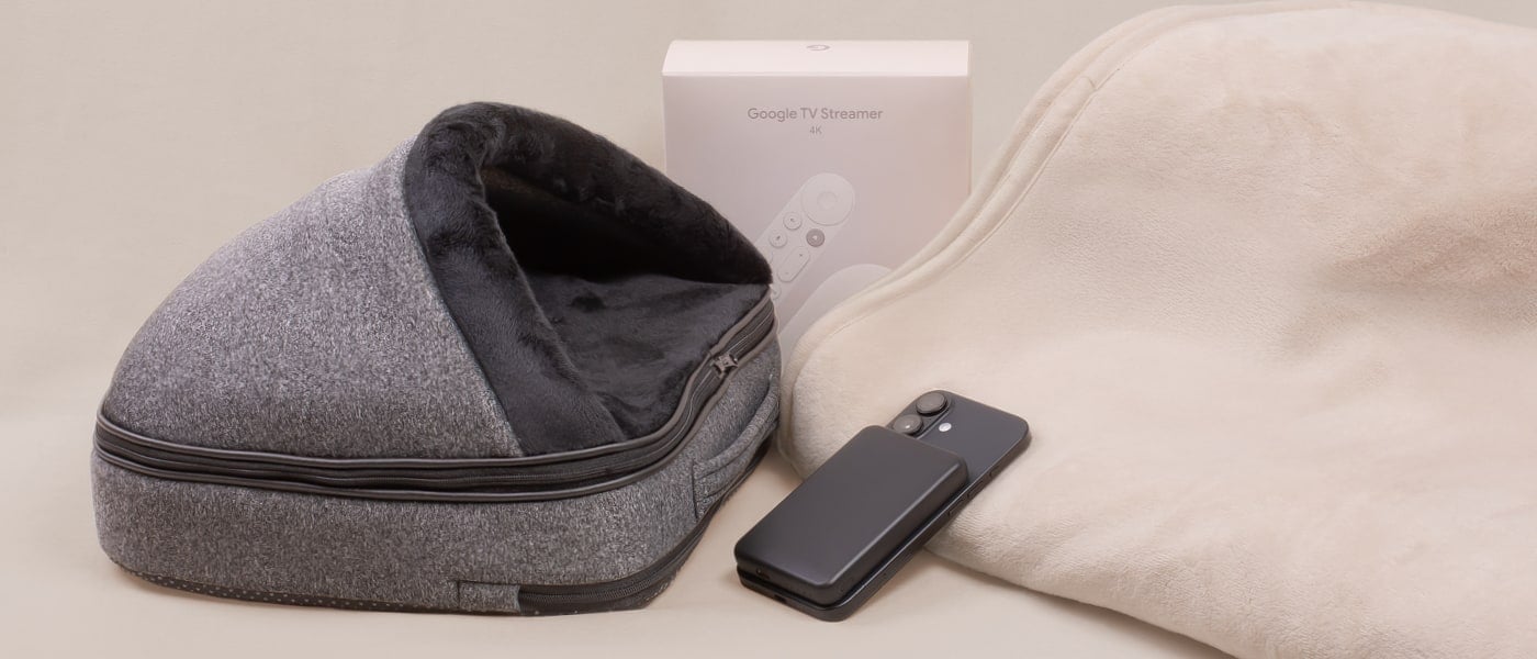 Grå fotvärmare med dragkedja och mjuk mörk insida på bord, bredvid beige kudde och fjärrkontroll. Förpackning med texten ”Google TV Streamer 4K” syns i bakgrunden.