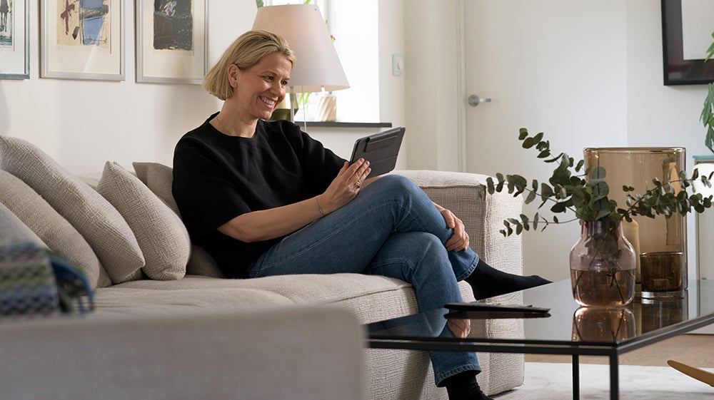 Smilende kvinne sitter i en sofa og bruker et nettbrett i en lys stue med moderne innredning.