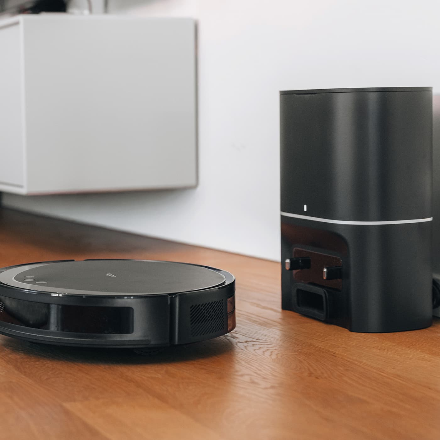 Svart robotdammsugare framför en automatisk tömningsstation på ett golv i trä.