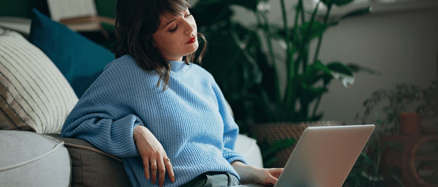 Kvinna i blå stickad tröja lutar sig mot en soffa och arbetar på en bärbar dator i ett rum med gröna växter.