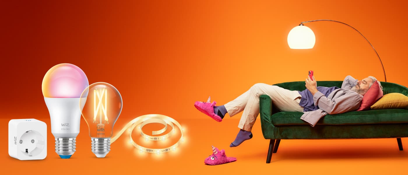 WiZ smartbelysning med lyspærer, LED-strip og kontakt; mann slapper av på grønn sofa med mobil i varm oransje setting.