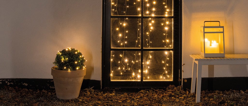 Utendørs pynt med varm hvit lyslenke i vindu, lys i potte og lykt på benk omgitt av høstløv.
