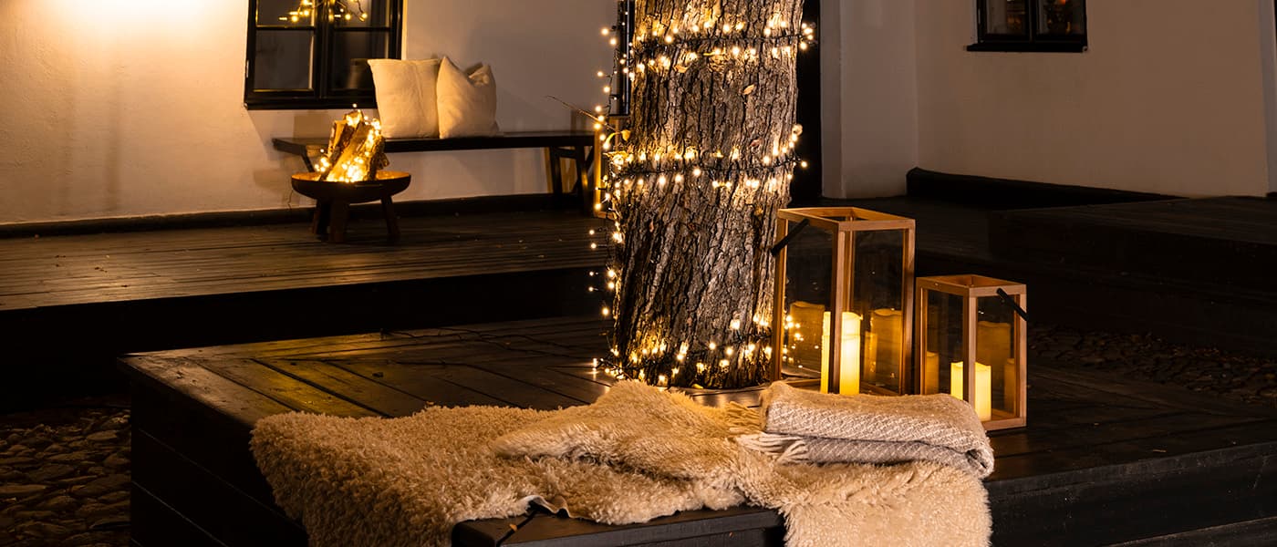 Upplyst trädstam med ljusslinga, tre lyktor med LED-ljus och filtar på en altan i kvällsbelysning.