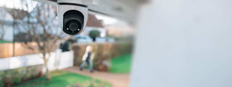 Overvåkningskamera montert under et tak, rettet mot en hagegang hvor en uklar person går forbi.