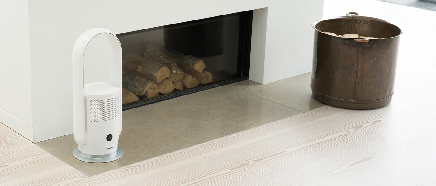 Hvit bladløs vifte med luftrenser plassert ved siden av en peis med stablet ved, ved siden av en kobberfarget vedbøtte.