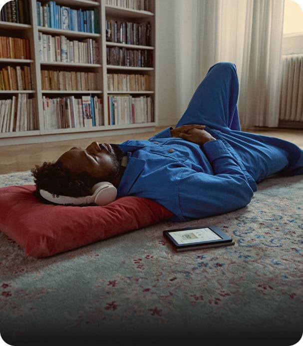 Man i blå kläder vilar på matta med hörlurar, bredvid en Kobo-läsplatta som visar ljudboksuppspelning.