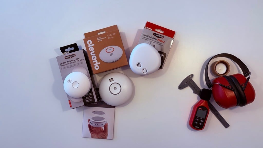 Tre røykvarslere med emballasje plassert på et hvitt bord, sammen med hørselsvern, et digitalt måleinstrument og en skyvelær.