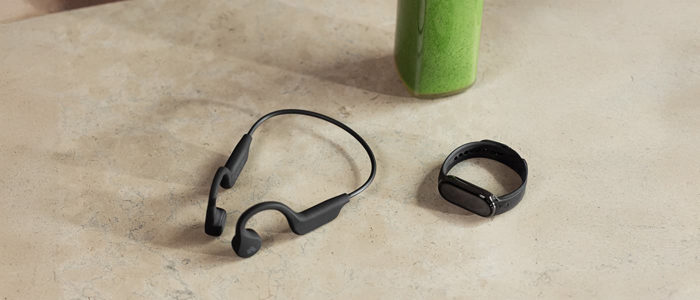 Svarta bone conduction-hörlurar och en svart aktivitetsklocka på ett bord, bredvid en grön smoothie.