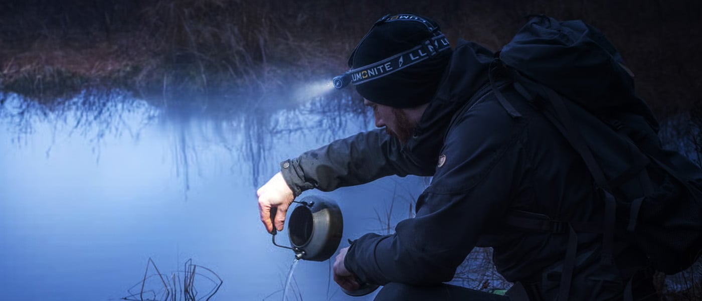 Pannlykt som lyser opp i mørket under friluftsliv. Mann heller vann ved en innsjø i skumringen.