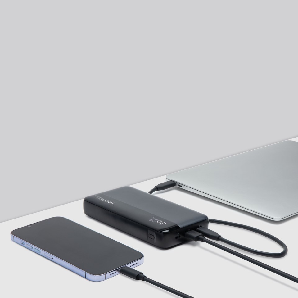 Svart powerbank laddar en bärbar dator och en smartphone via USB-C-kablar på ett vitt bord.