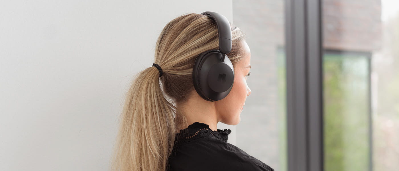 Kvinna med hästsvans bär svarta over-ear-hörlurar och tittar mot ett fönster.