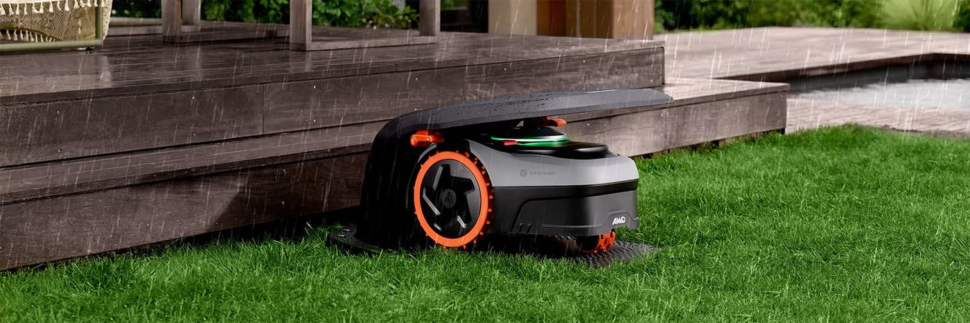 Segway robotgräsklippare med AWD och orange hjul parkerad i sitt garage på gräsmattan i regn intill ett trädäck."