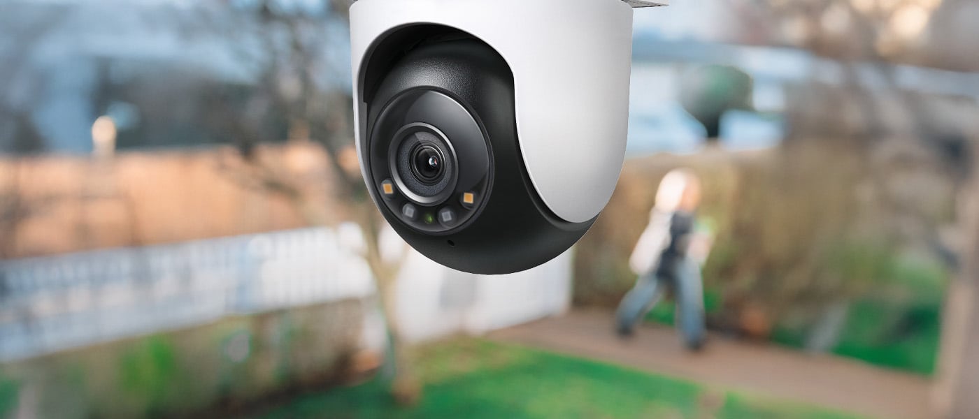 Nærbilde av et hvitt og svart utendørs overvåkingskamera montert under tak, med uskarp hage og person i bakgrunnen.