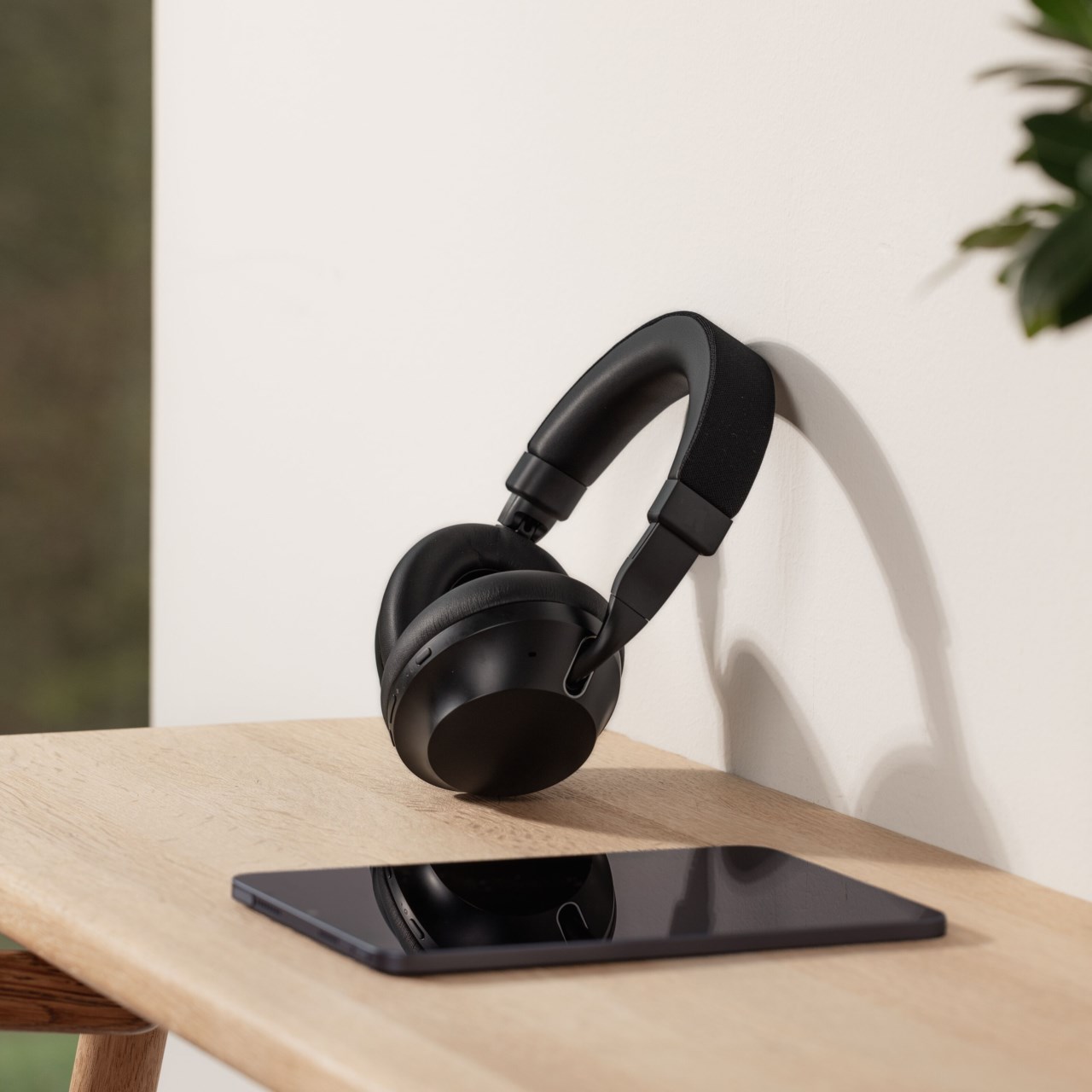 Svarta over-ear-hörlurar hänger på en vit vägg ovanför ett träbord med en svart surfplatta.