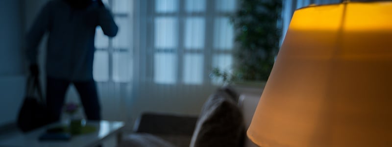 Innbruddstyv i et mørklagt rom i bakgrunnen, med en tent gulvlampe i fokus foran.