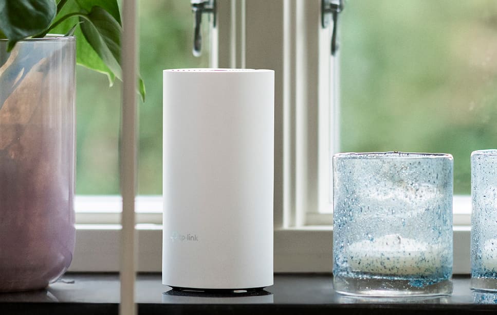 Vit TP-Link-router placerad i ett fönster, mellan en krukväxt och två ljuslyktor i glas.