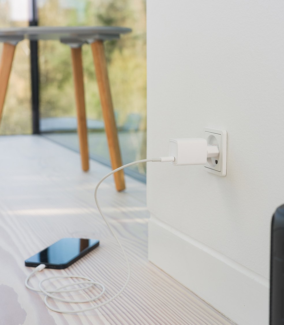 Smartphone laddas med vit väggladdare och kabel i eluttag nära golvet.
