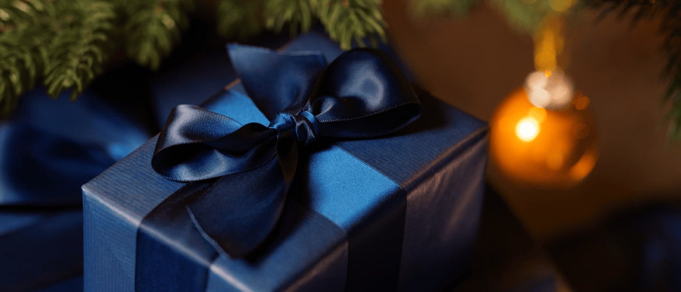 Julegave pakket inn i mørkeblått papir med blå silkebånd, granbar og en gullfarget julekule i bakgrunnen.
