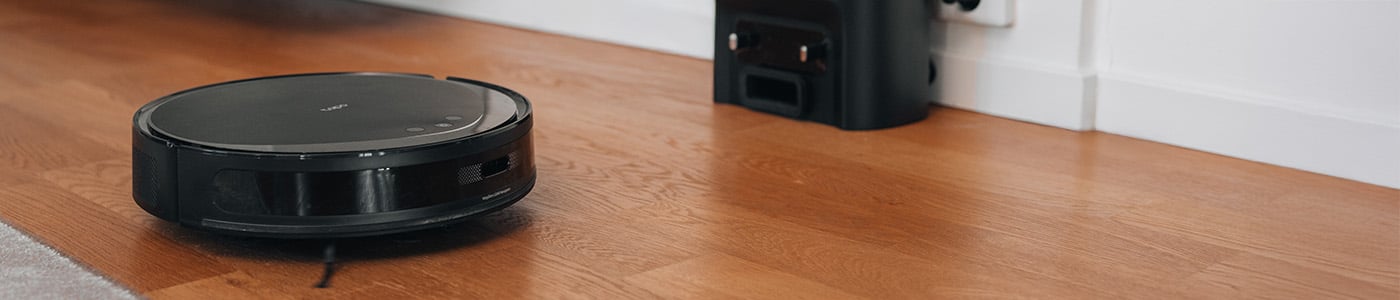 Svart robotdammsugare på trägolv, på väg mot sin laddstation vid vit vägg.