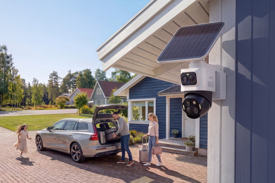 Eufy overvåkningskamera med solcellepanel montert på husvegg, familie laster av bil i bakgrunnen.