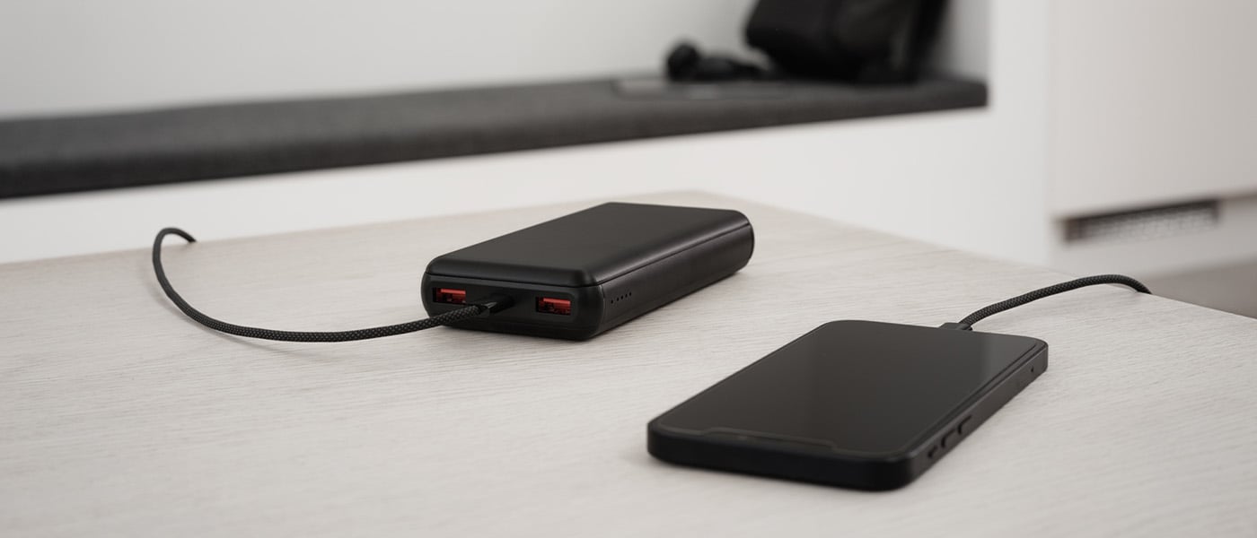 Svart powerbank med två USB-portar laddar en smartphone via svart textilklädd kabel på ett ljust bord i modern miljö.