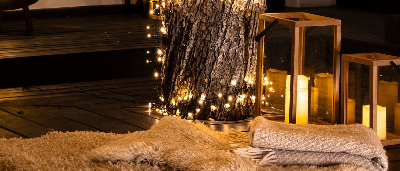 Upplyst träd med varmvita ljusslingor, lyktor med LED-ljus och vikta filtar på ett trädäck i kvällsmiljö.