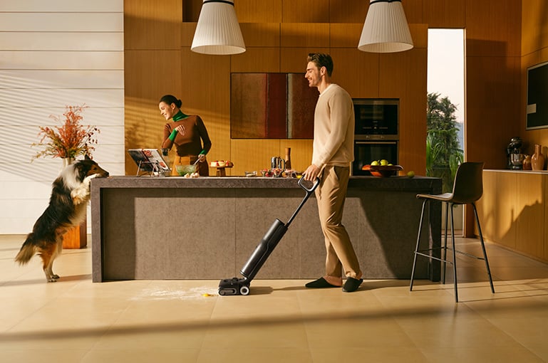 Man städar upp spill på golvet med en svart handdammsugare i ett modernt kök, hund och kvinna i bakgrunden.