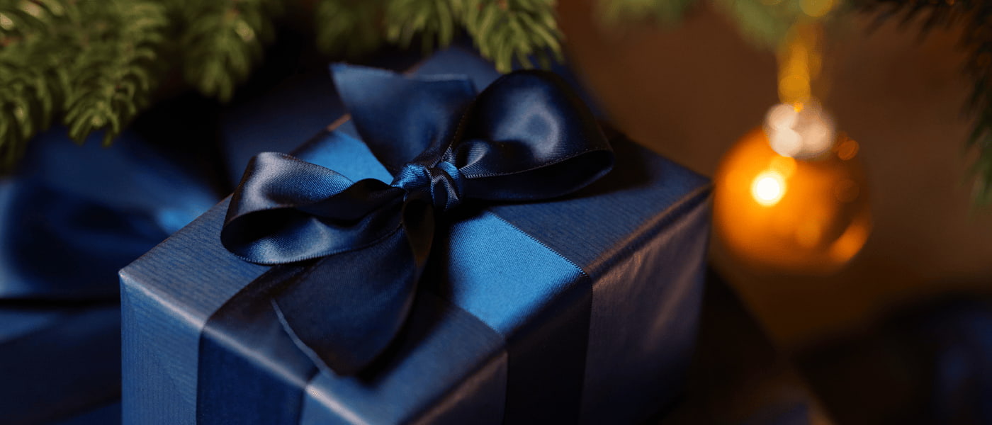Julklapp inslagen i mörkblått papper med blå sidenrosett, granris och gul julkula i bakgrunden.