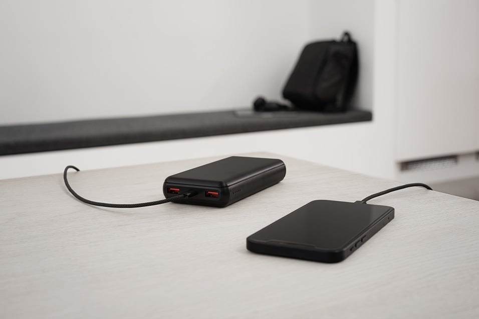 Svart powerbank med to USB-porter lader en smarttelefon via svart stoffbelagt kabel på et lyst bord i moderne rom.