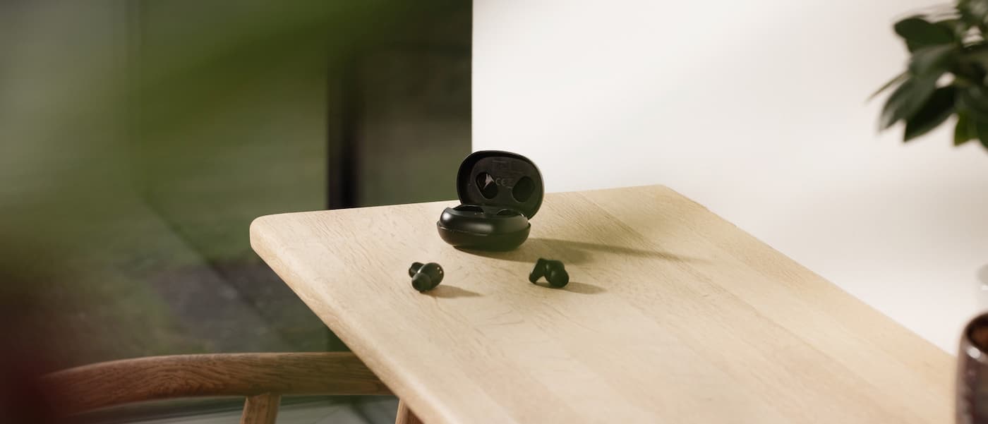 Svarte trådløse in-ear-hodetelefoner med åpent ladeetui, plassert på et lyst trebord i et minimalistisk innemiljø.