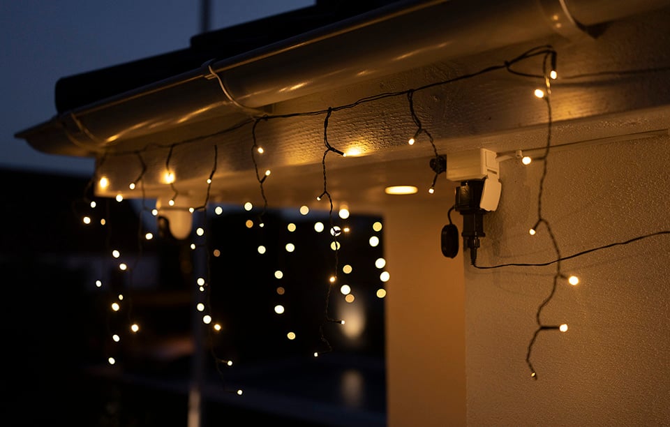 Varmvit ljusslinga med hängande ljuspunkter monterad under takkant på husfasad i kvällsljus.
