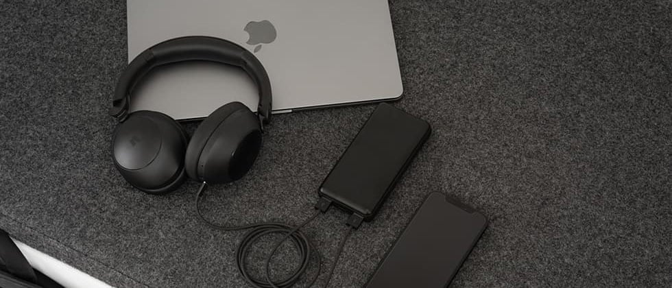 Bärbar dator, svarta over-ear-hörlurar, powerbank och smartphone på grå filtduk, allt med kablar anslutna.