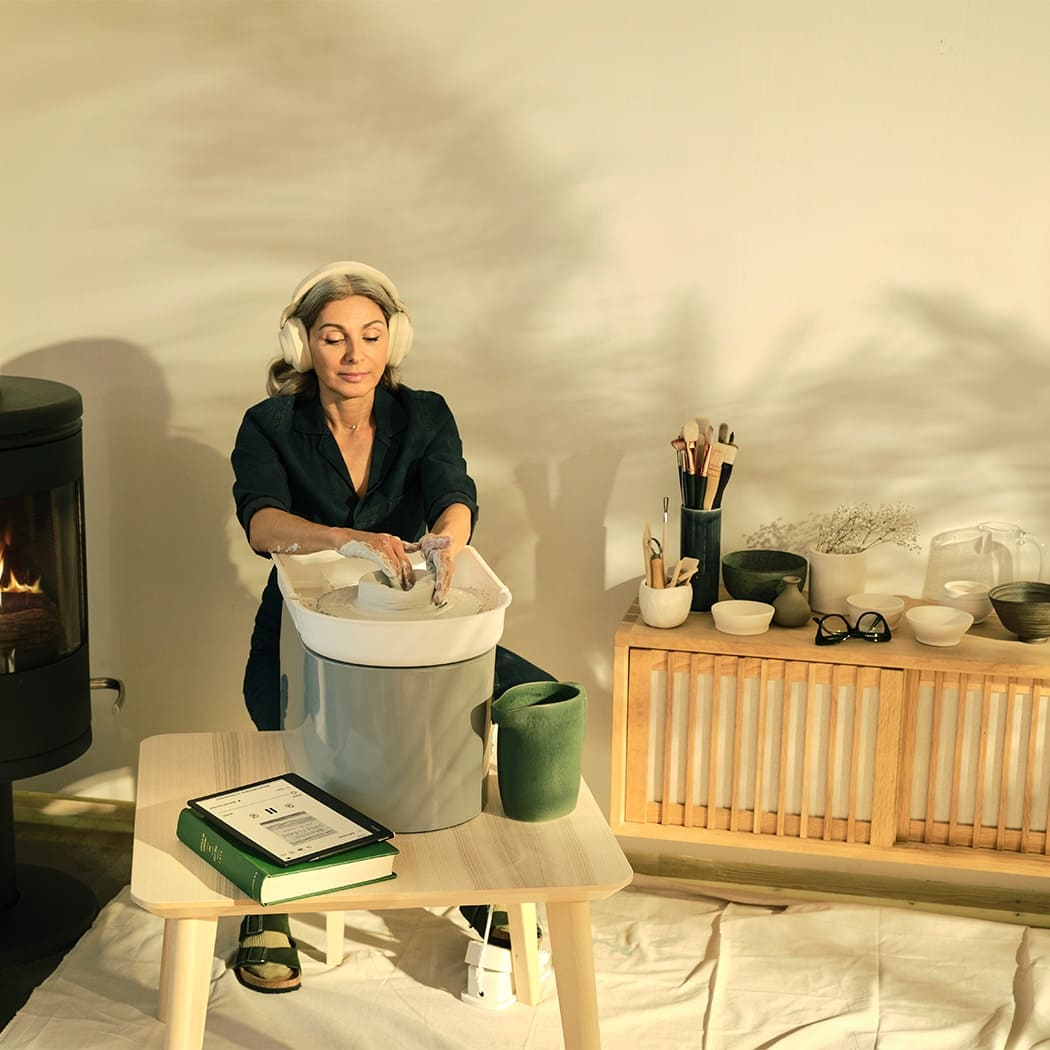 Kvinna med hörlurar drejar vid en lerkruka, med en Kobo-läsplatta på bordet bredvid en bok, i varm hemmamiljö.