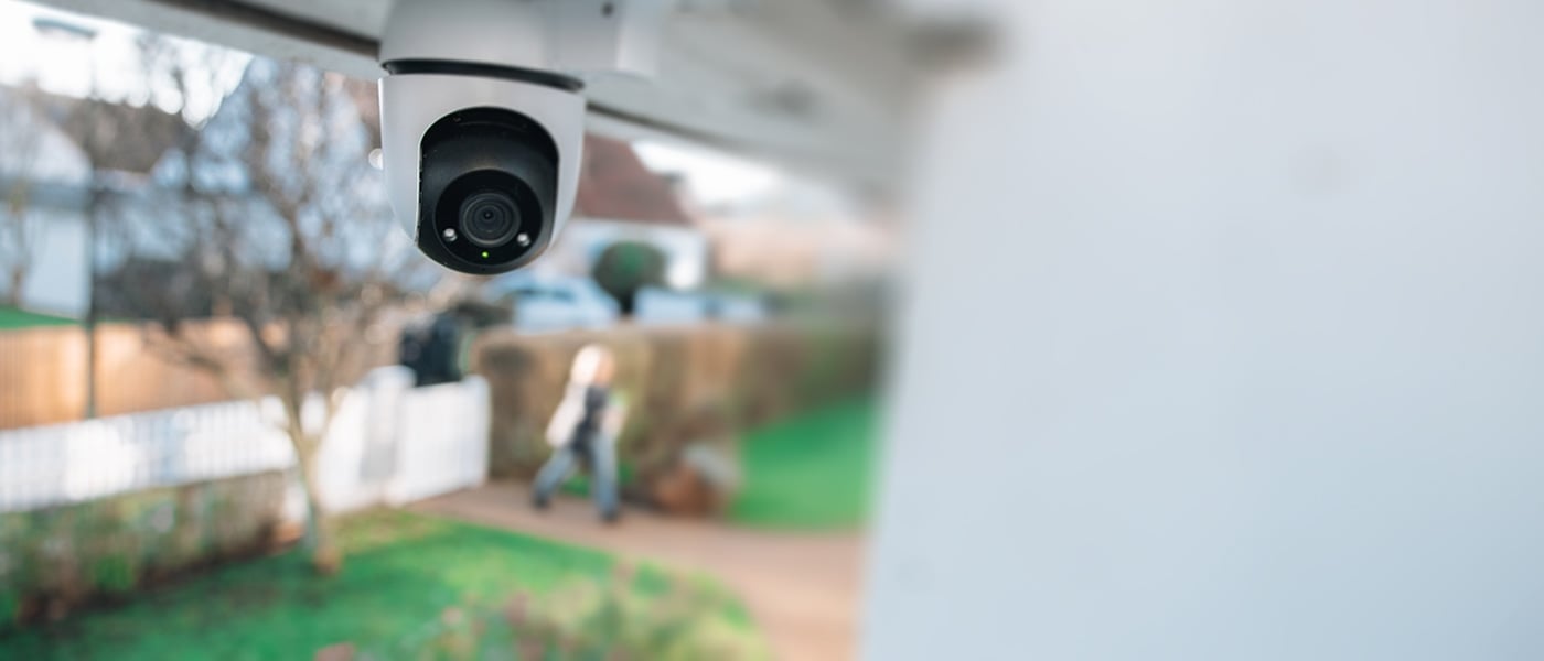 Overvåkningskamera montert under et tak, rettet mot en hagegang der en uklar person passerer.