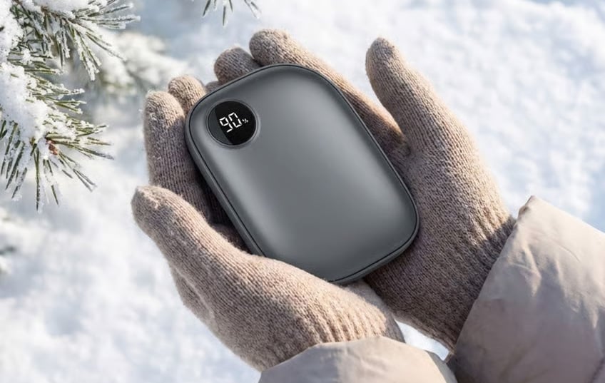 Person med stickade vantar håller i en svart handvärmare med digital display som visar 90 %. Snö och gran i bakgrunden.